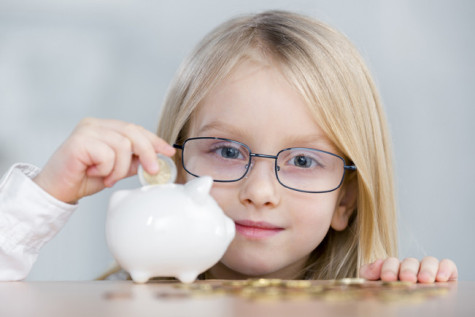 Cute little girl wearing eyeglasses putting coin into piggy bank